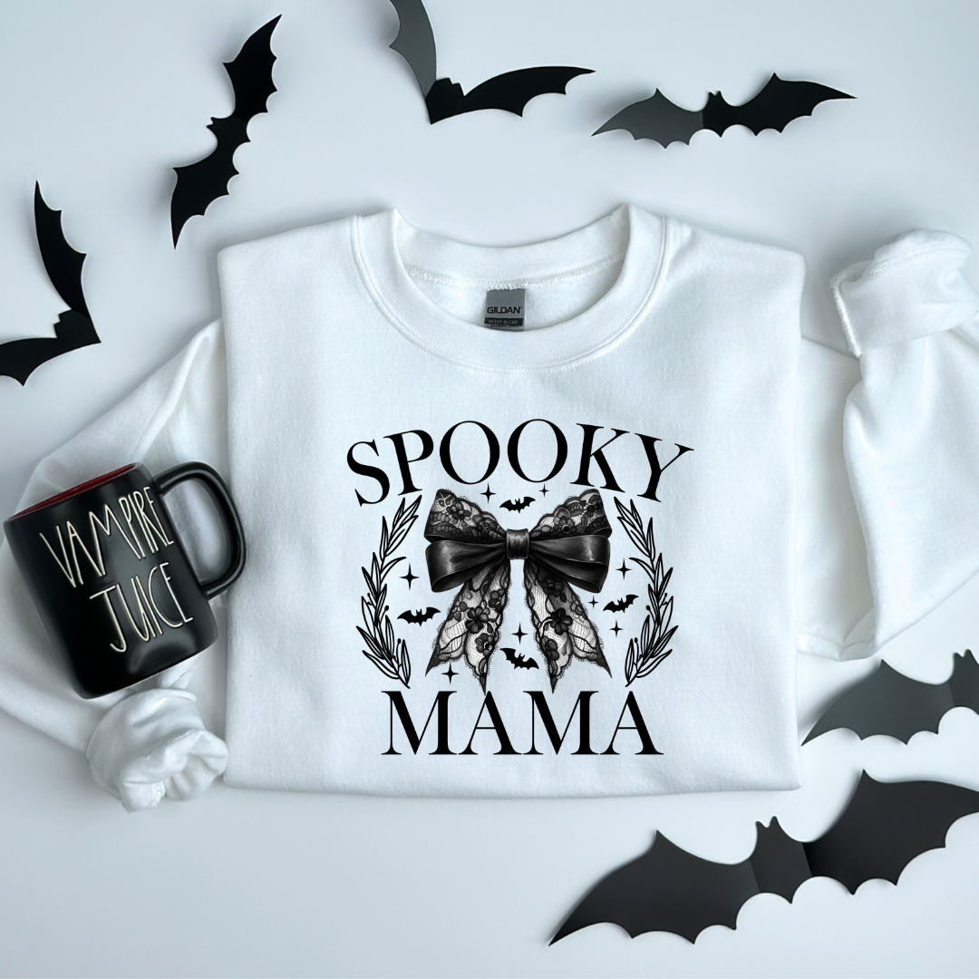 A white crewneck sweatshirt with 'Spooky Mama' text and a design featuring a black bow, bats, and a wreath, presented on a surface with bat cut-outs and a mug with 'BAT JUICE' text.