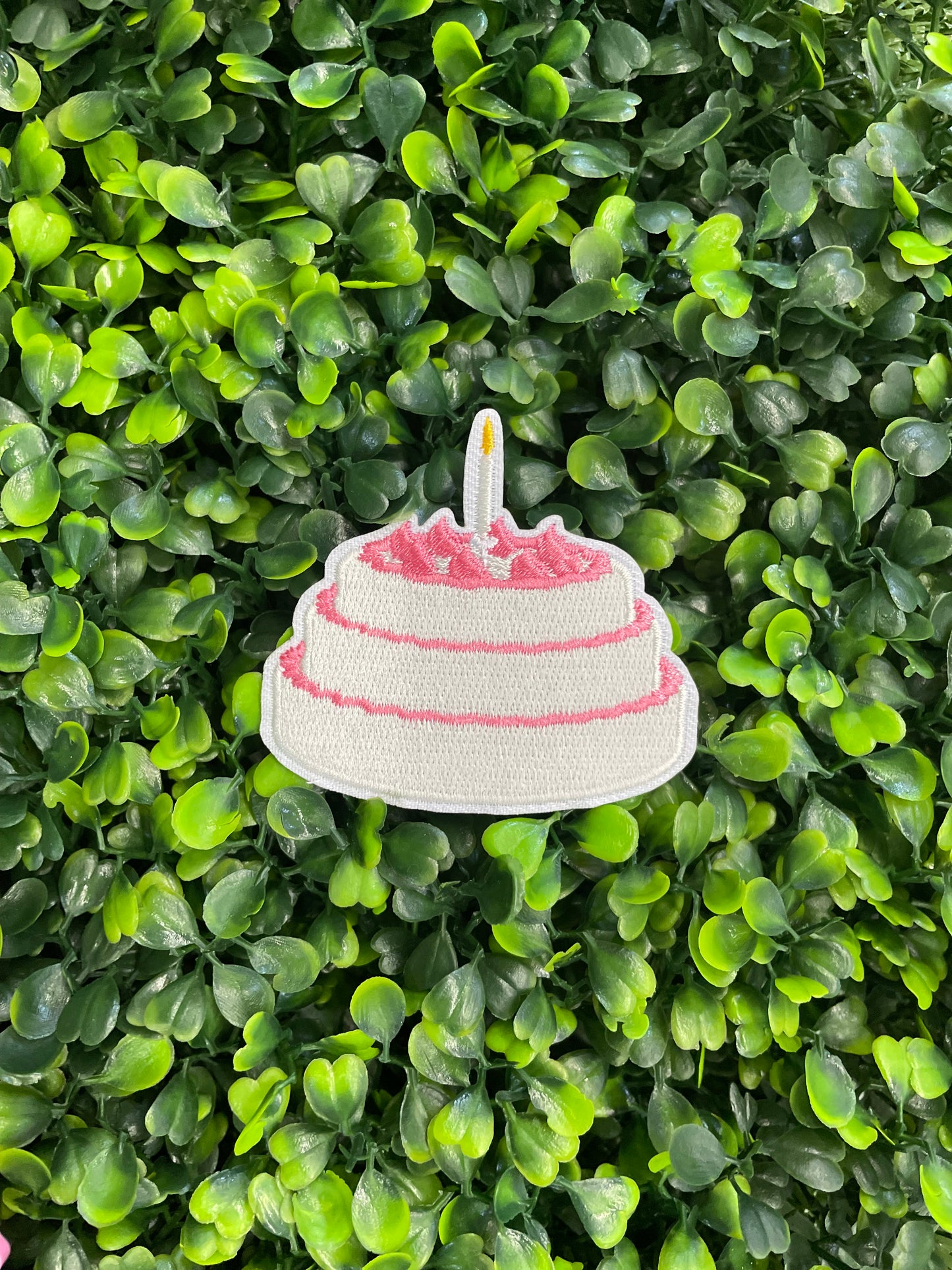 A small pink and white birthday cake embroidered patch placed on top of a green leafy background