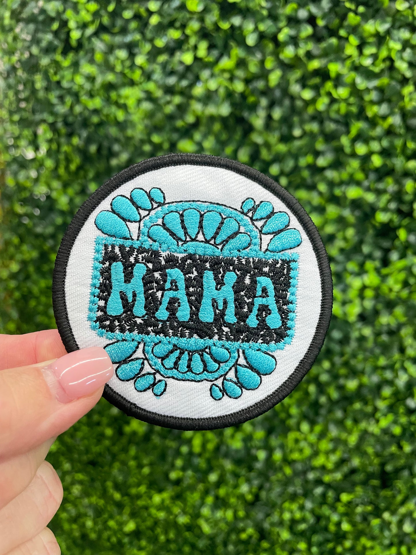 Circular patch with 'MAMA' text and teal designs held against a green textured background