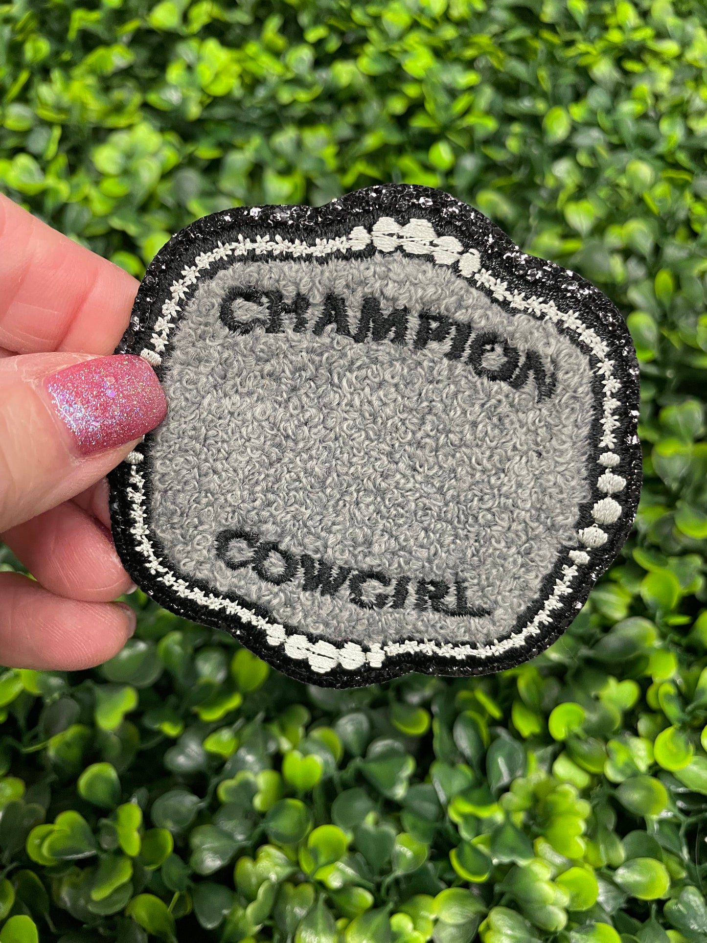 A close-up of an embroidered patch with the text 'Champion Cowgirl' held against a green leafy background.