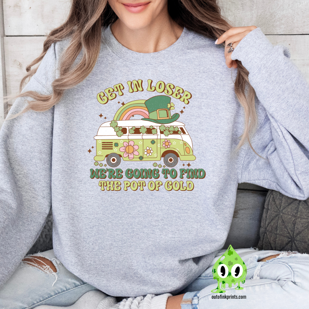 A woman wearing a grey sweatshirt with a St. Patrick's Day theme, featuring text and a graphic of a rainbow, clover, and what appears to be a leprechaun.