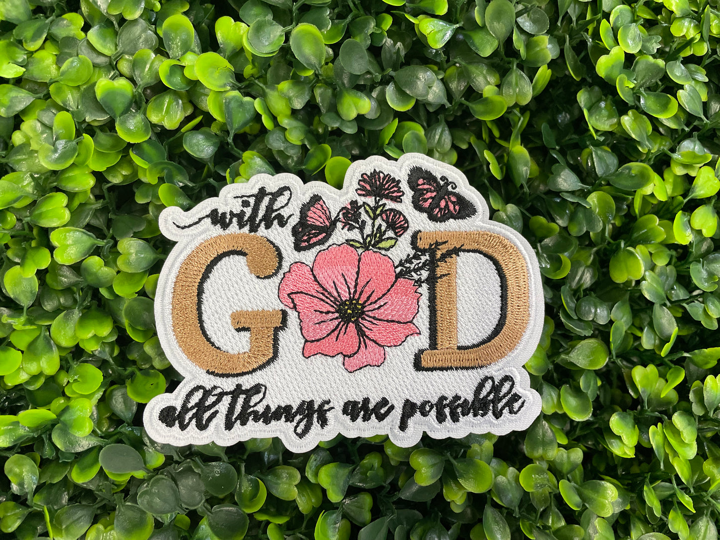 Embroidered patch butterflies and flowers and text stating "with God all things are possible", held against a green leafy background