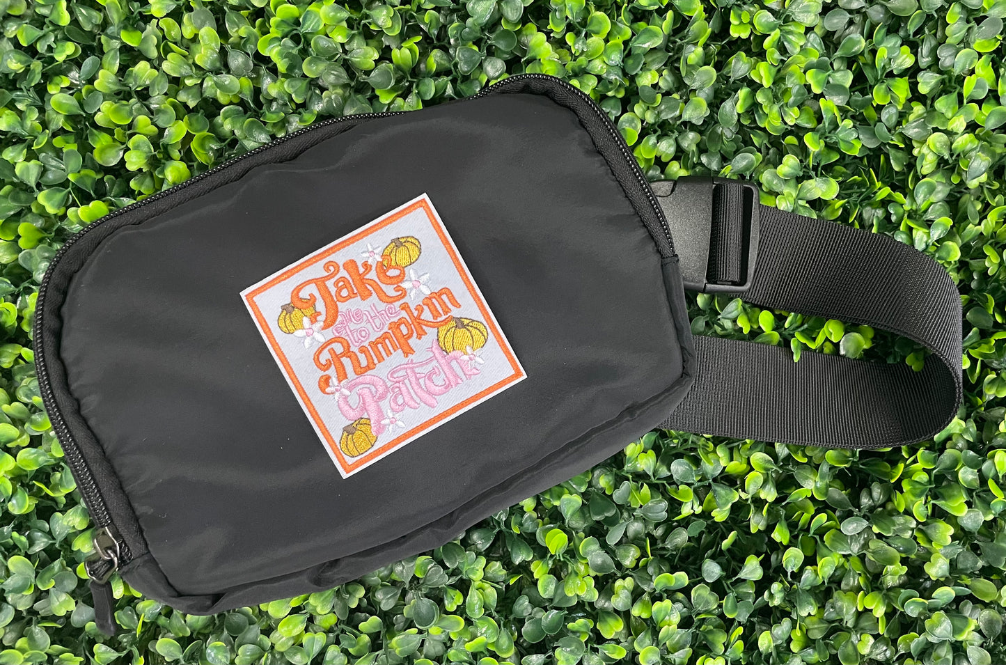Embroidered patch featuring orange and pink text "Take me to the pumpkin patch" with pumpkin and flower designs around it, placed on a black fanny pack