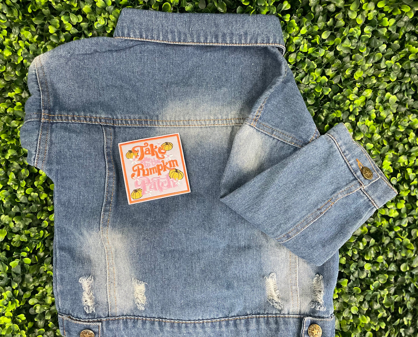 Embroidered patch featuring orange and pink text "Take me to the pumpkin patch" with pumpkin and flower designs around it, placed on a children's denim jean jacket