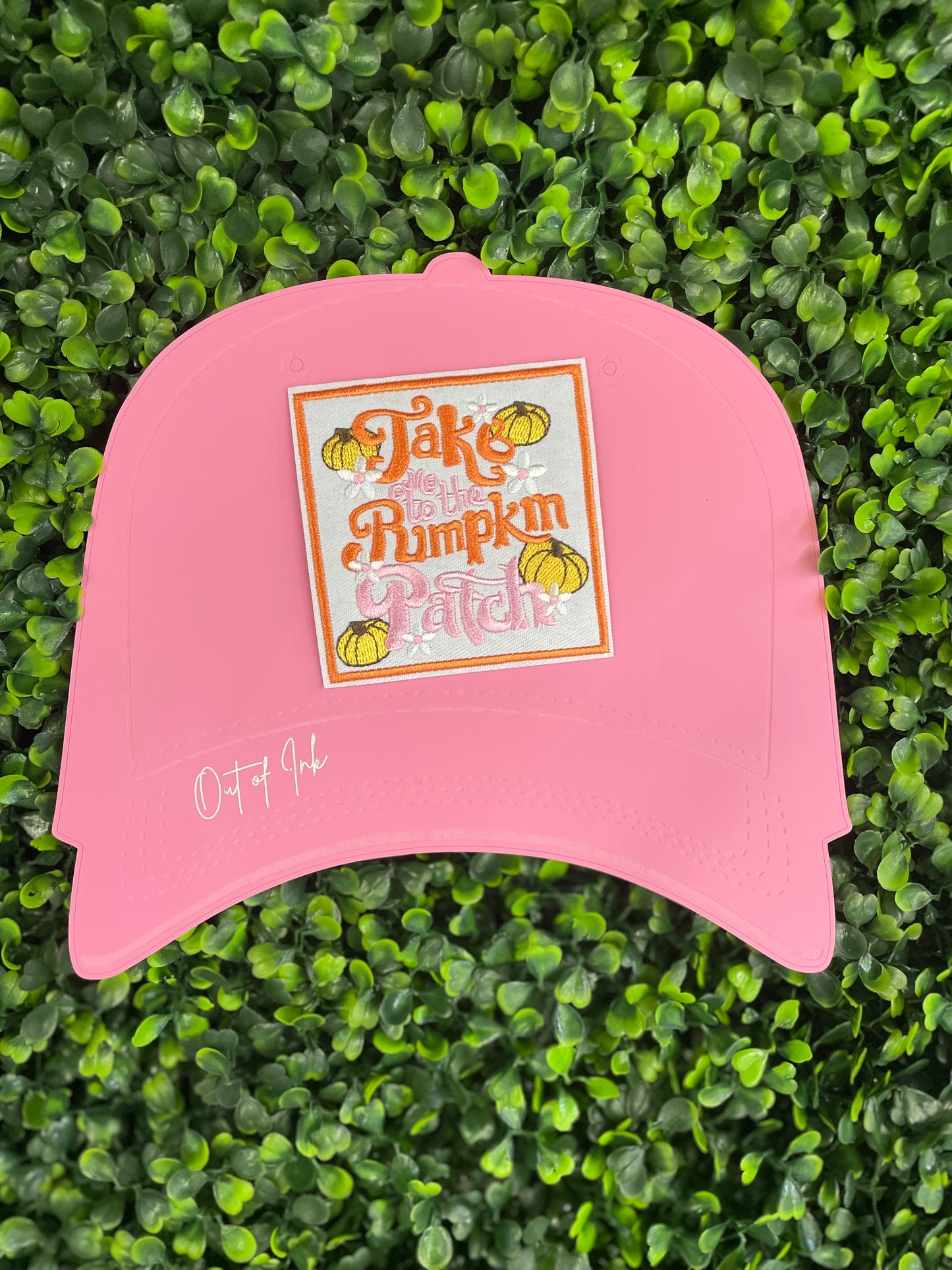 Embroidered patch featuring orange and pink text "Take me to the pumpkin patch" with pumpkin and flower designs around it, placed on a pink trucker hat graphic