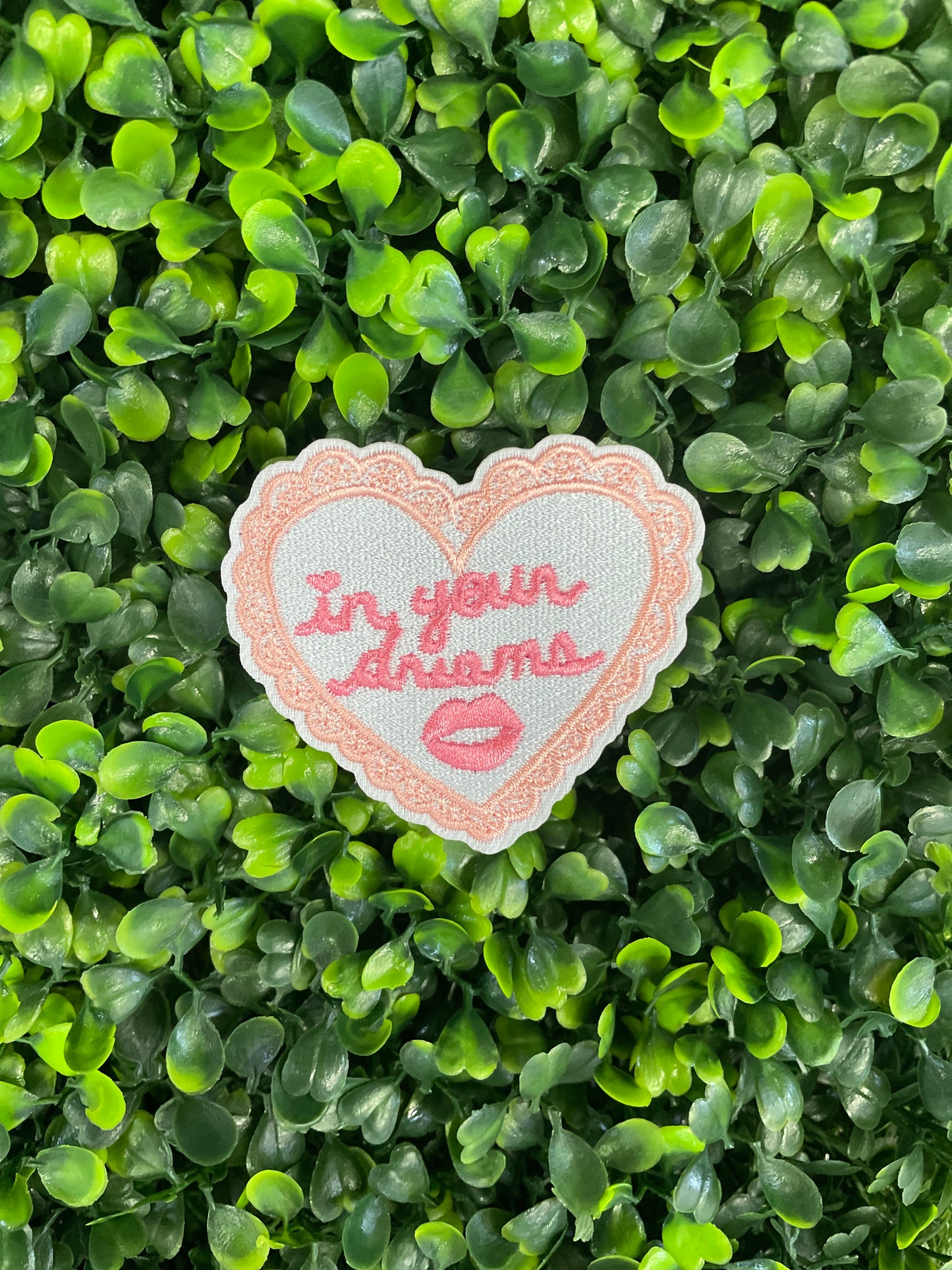 Heart-shaped embroidered patch with 'In your dreams' text held against a green and leafy background