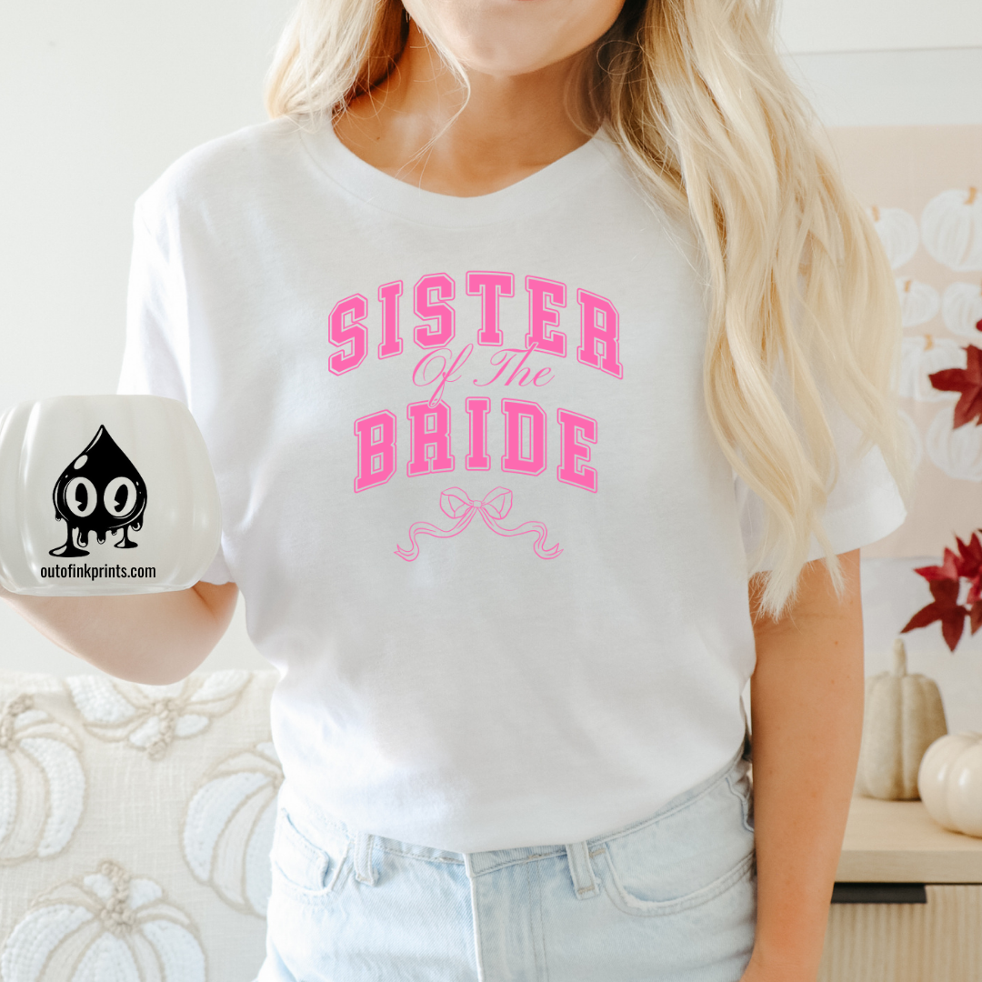 A woman sitting wearing a white t-shirt with the text 'Sister of the Bride' and a pink bow design on it.