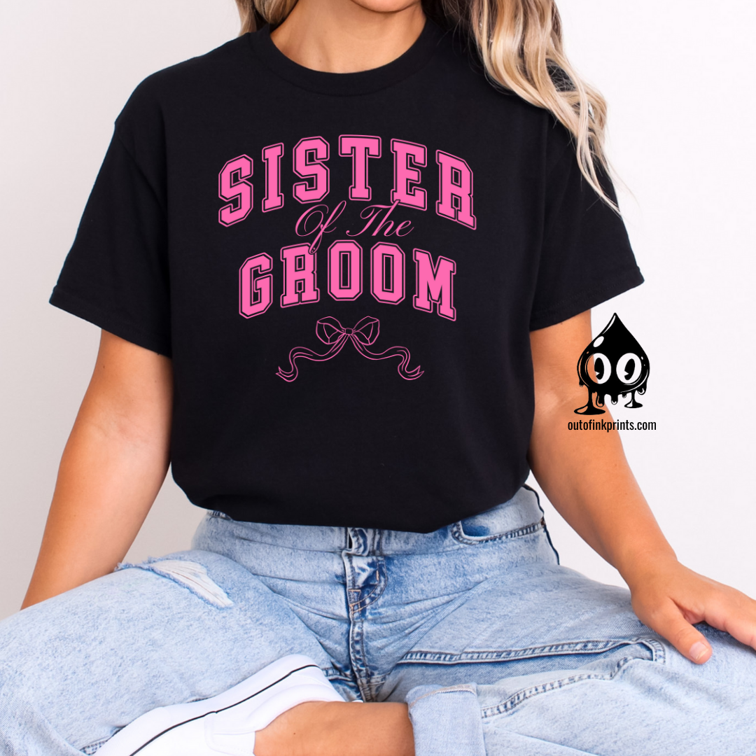 A woman sitting wearing a black t-shirt with the text 'Sister of the Groom' and a pink bow design on the front.