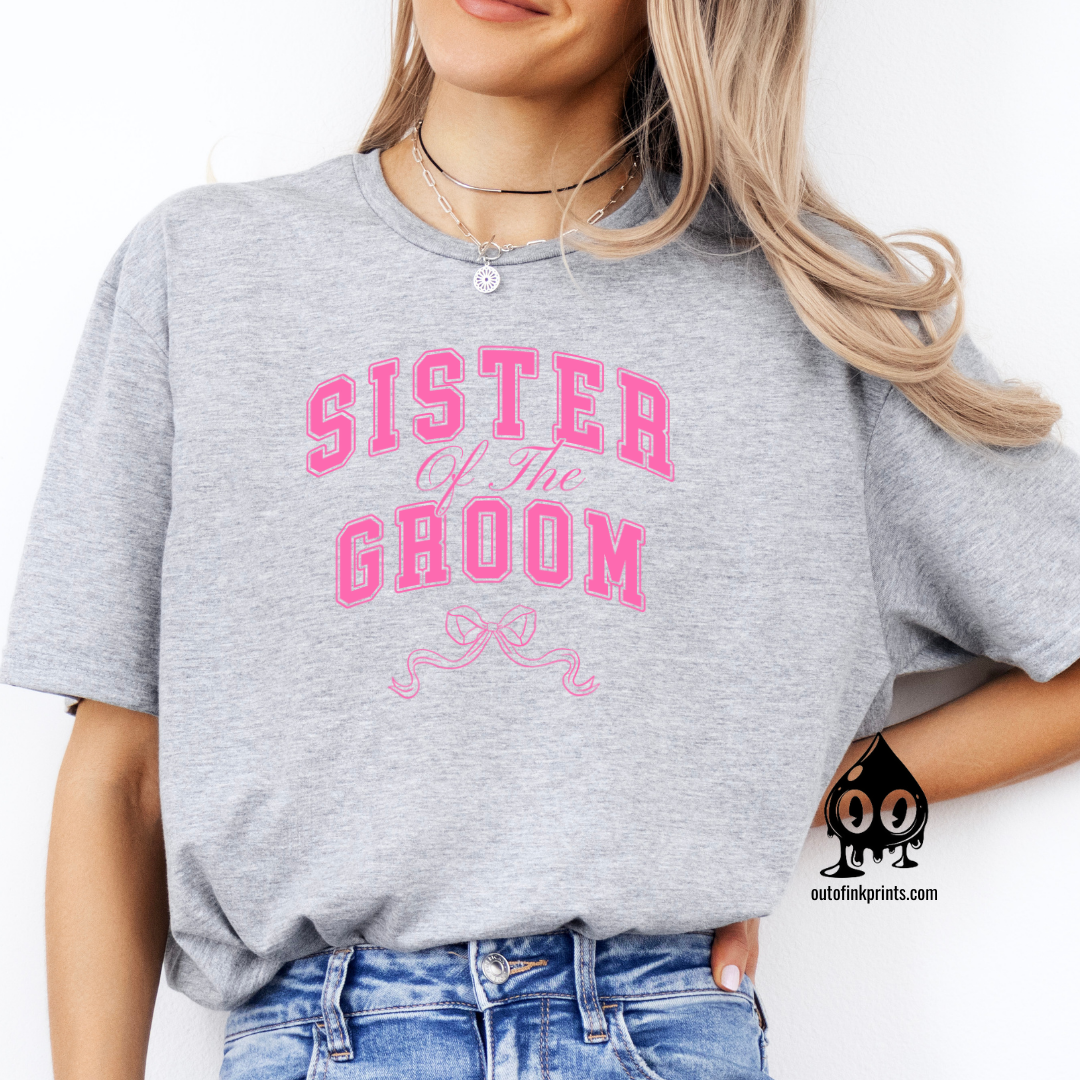 A woman sitting wearing a gray t-shirt with the text 'Sister of the Groom' and a pink bow design on the front.