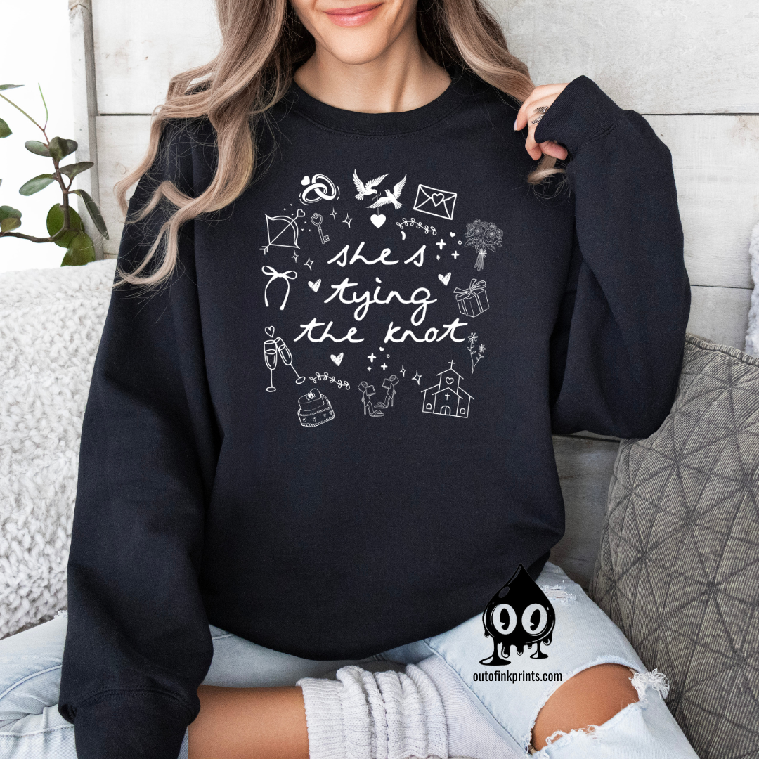 A woman smiling, wearing a black sweatshirt with white text and graphics related to a bride tying the knot.