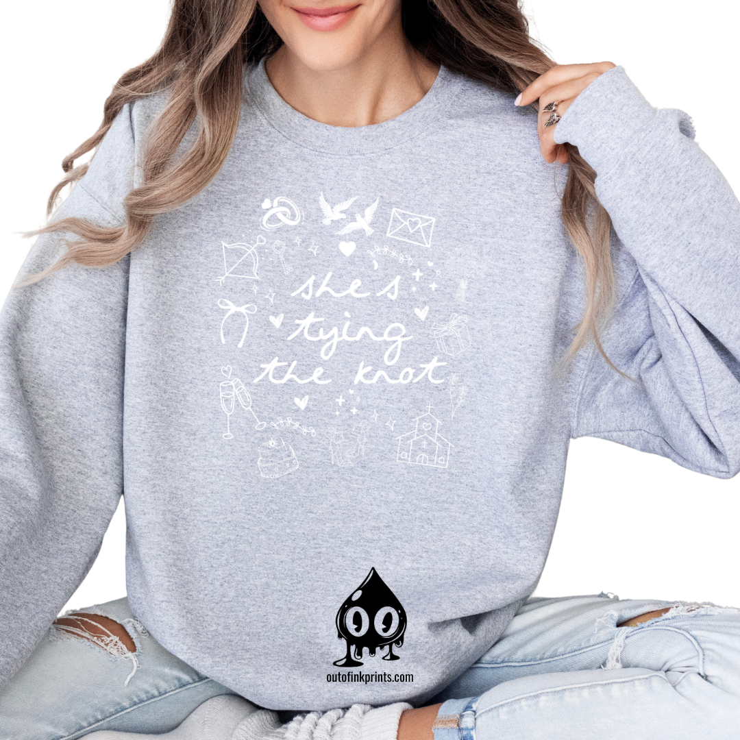 A woman smiling, wearing a gray sweatshirt with white text and graphics related to a bride tying the knot.