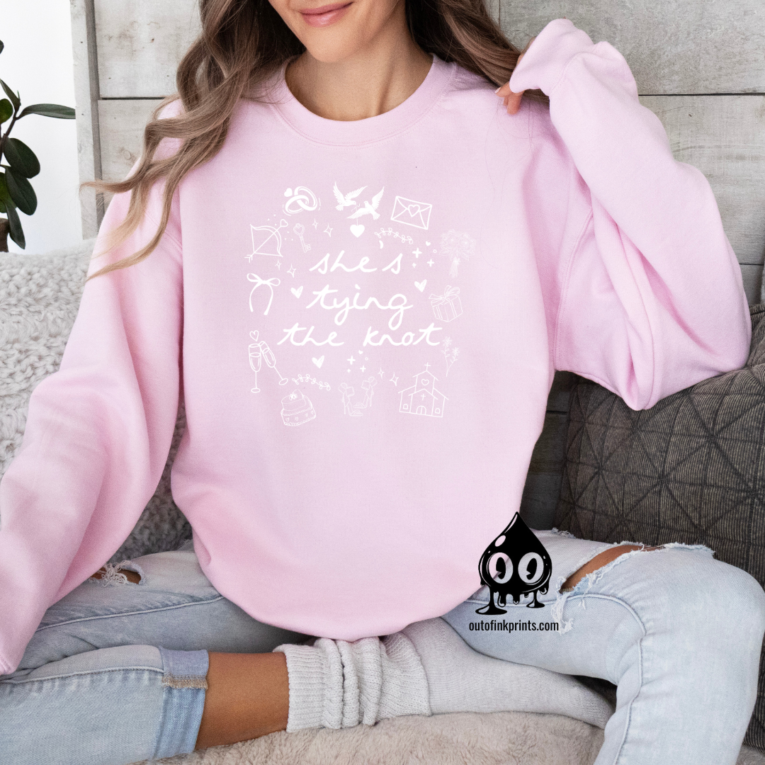 A woman smiling, wearing a light pink sweatshirt with white text and graphics related to a bride tying the knot.