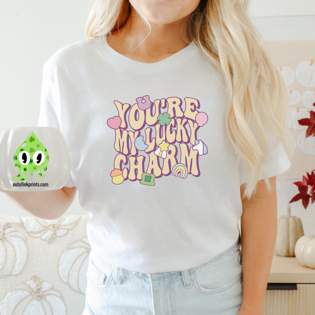 Front view of a white t-shirt with the text 'YOU'RE MY LUCKY CHARM' in a playful font, surrounded by decorative elements like shamrocks and hearts.