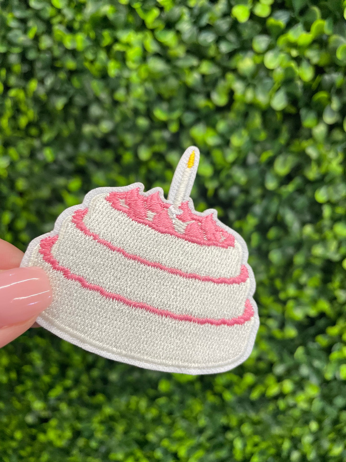 A small pink and white birthday cake embroidered patch placed on top of a green and leafy background