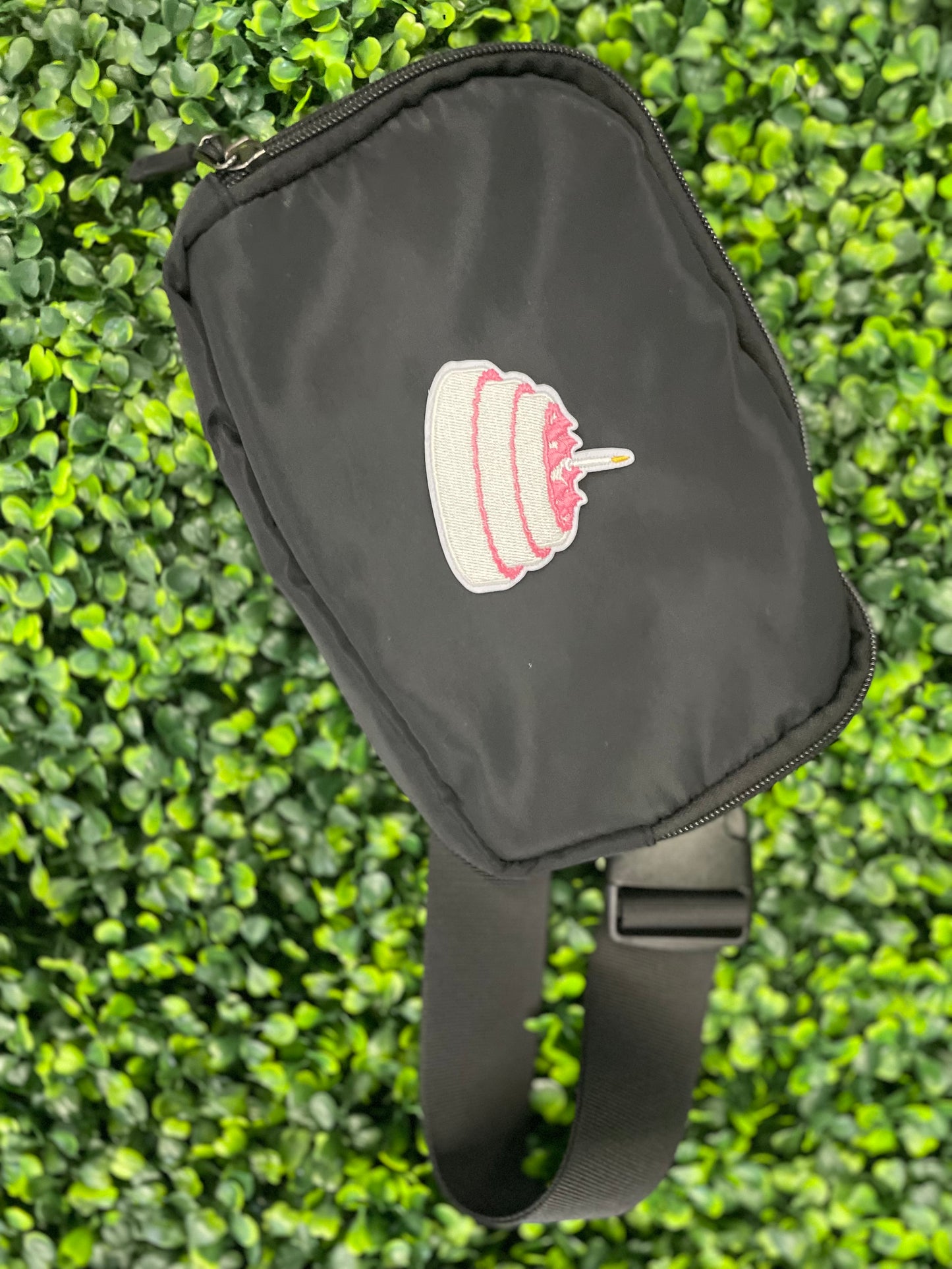 A small pink and white birthday cake embroidered patch placed on top of a small black bag