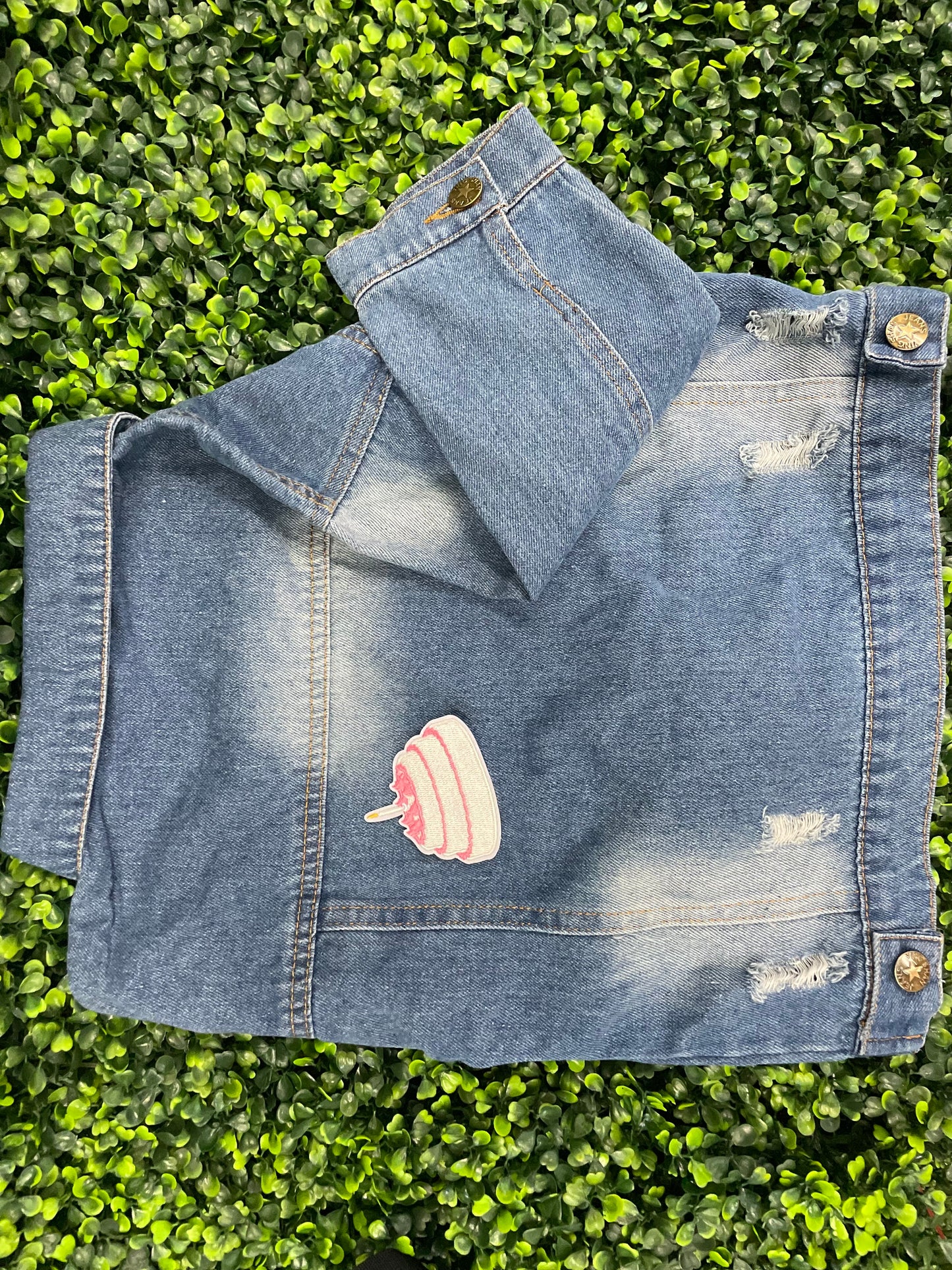 A small pink and white birthday cake embroidered patch placed on top of a children's denim jean jacket