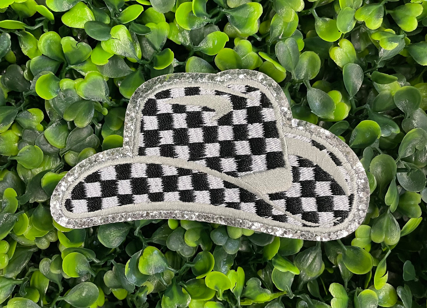 Checkered white and black cowboy hat patch with silver lining, placed onto a leafy backdrop