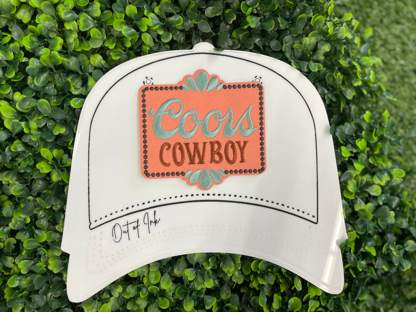 Embroidered patch with 'Coors Cowboy' text on a light orange background with teal accents, placed on a white trucker hat graphic