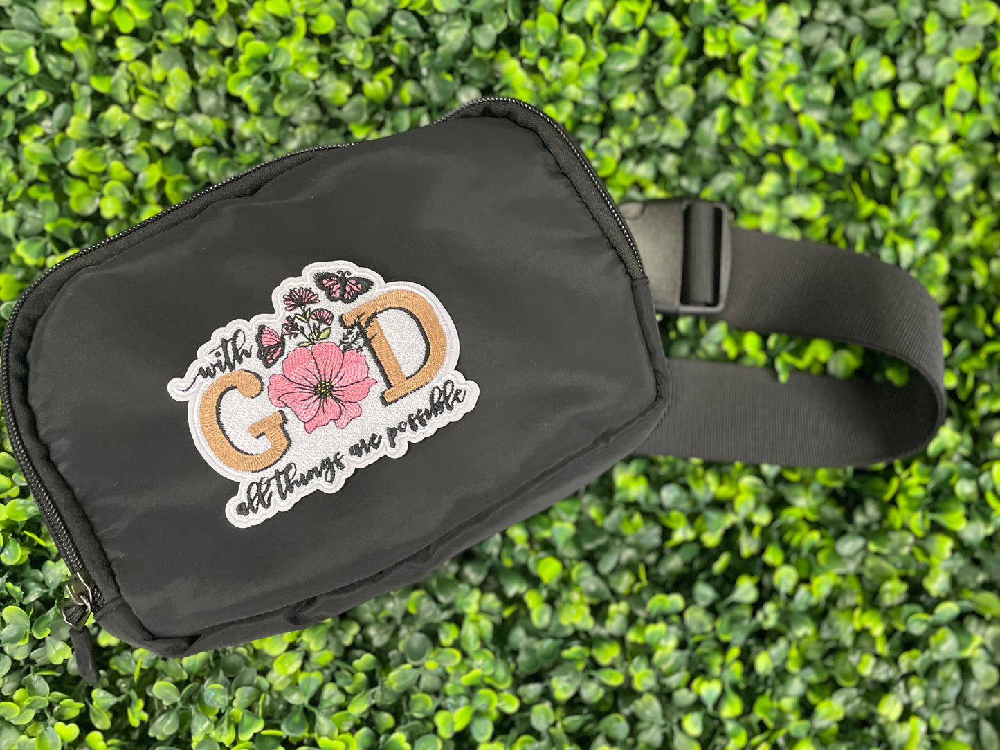 Embroidered patch butterflies and flowers and text stating "with God all things are possible", held against a black fanny pack