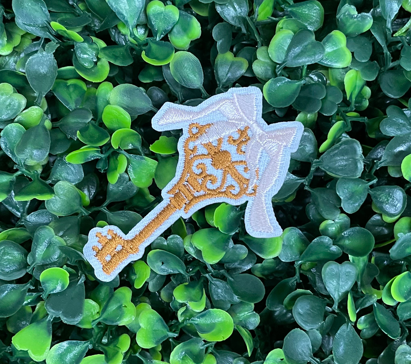 A patch in the shape of a vintage key with a gold bow, embroidery work, set on a background of greenery