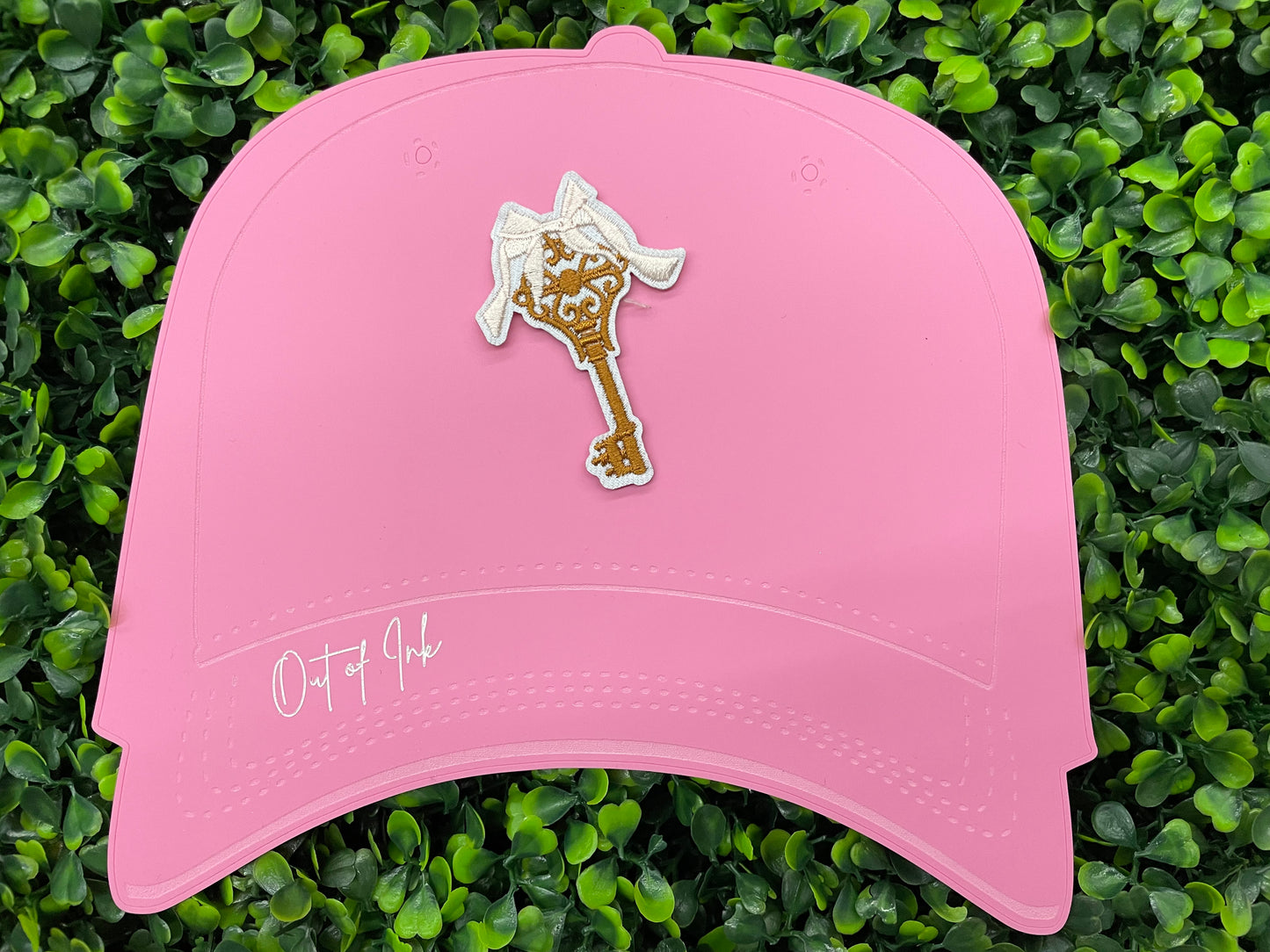 A patch in the shape of a vintage key with a gold bow, embroidery work, on a pink hat against a greenery background