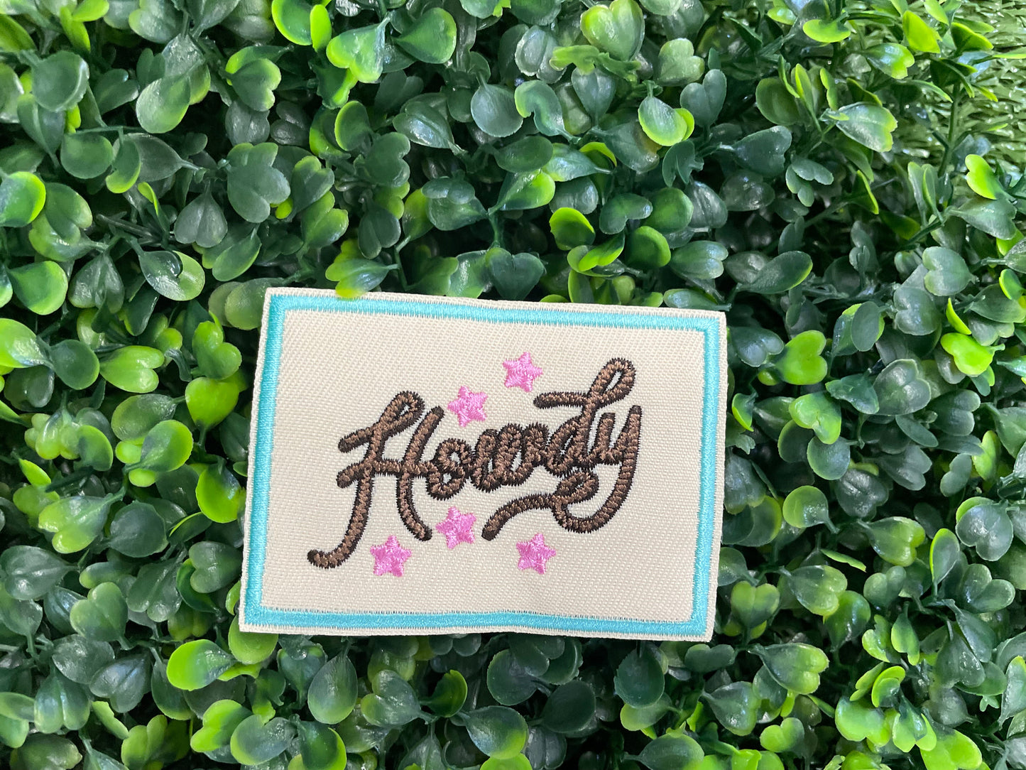 Embroidered patch with brown "Howdy" text on a tan background with a light blue border, placed against a green leafy background