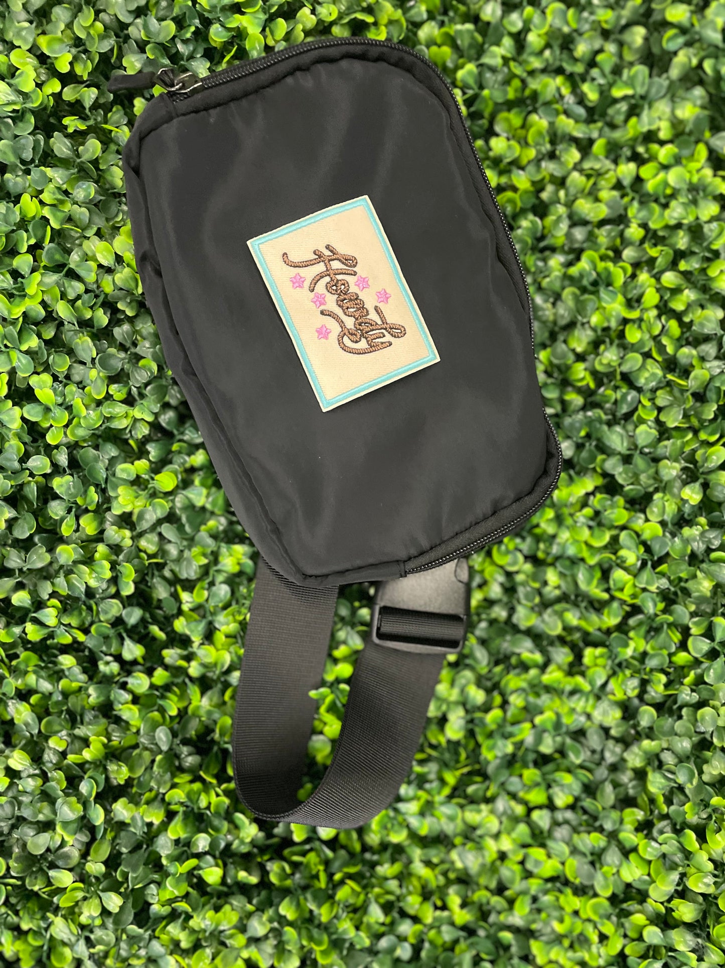Embroidered patch with brown "Howdy" text on a tan background with a light blue border, placed against a black fanny pack