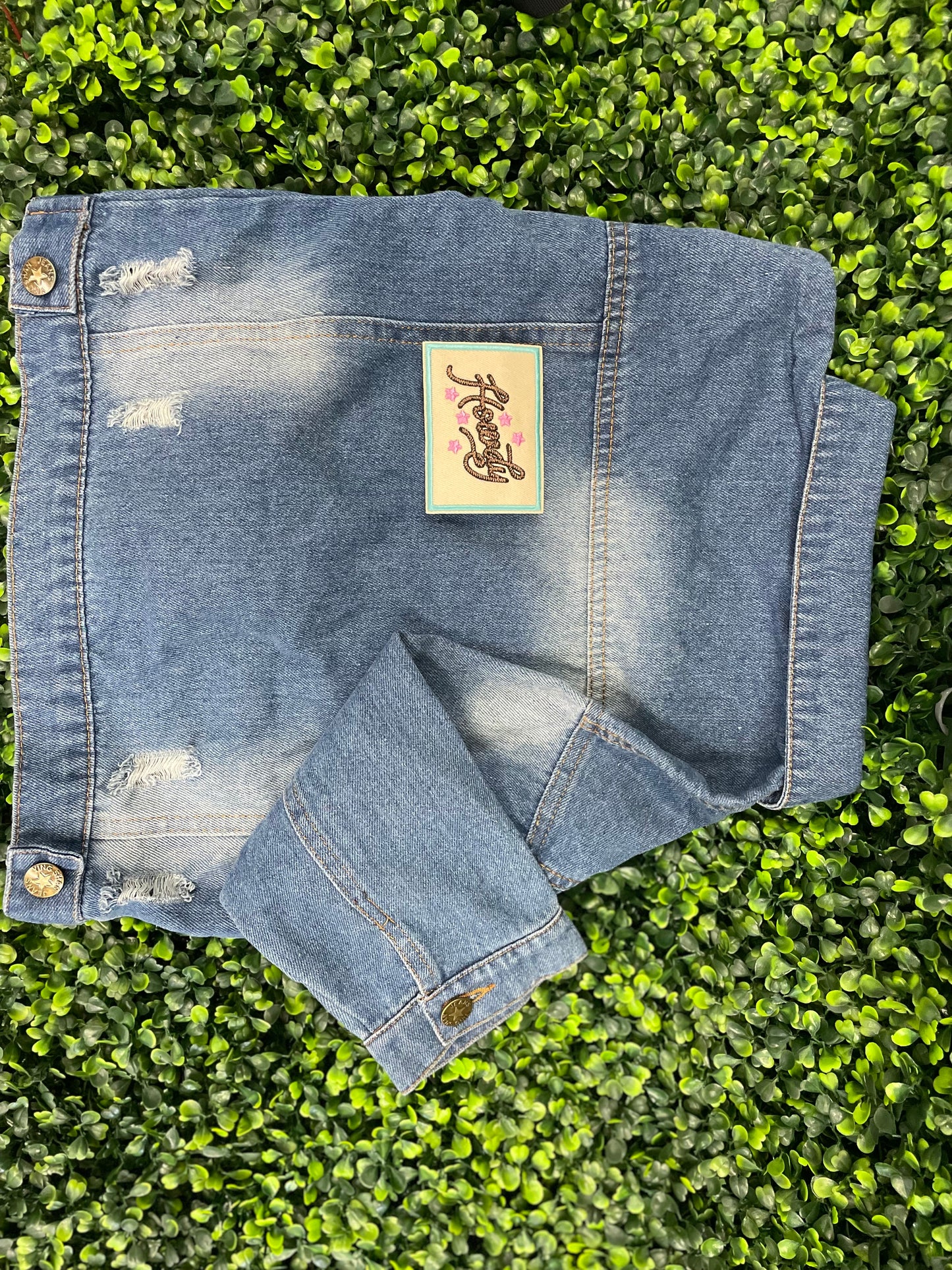Embroidered patch with brown "Howdy" text on a tan background with a light blue border, placed on a children's denim jean jacket