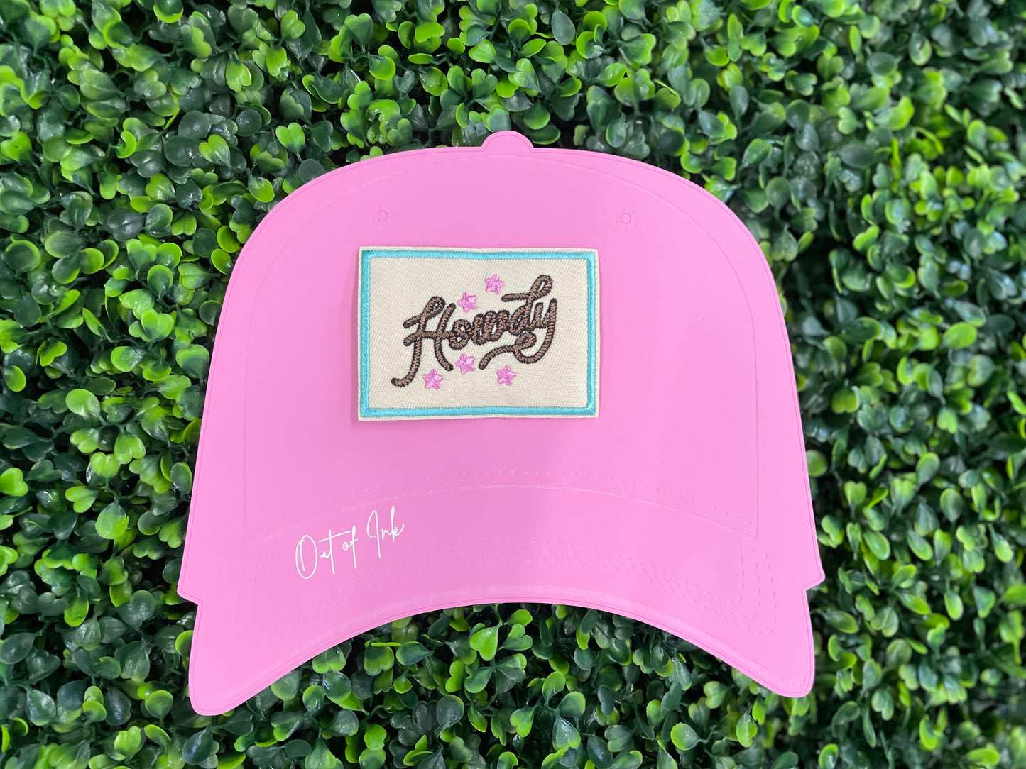 Embroidered patch with brown "Howdy" text on a tan background with a light blue border, placed against a pink trucker hat graphic