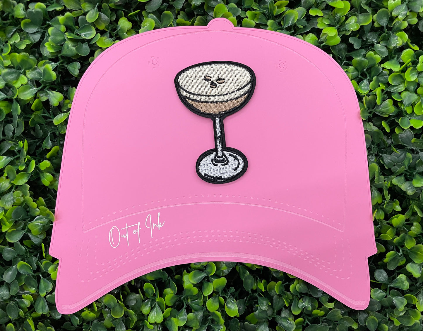 An iron on embroidered martini glass depiction set on a pink hat against a green backdrop