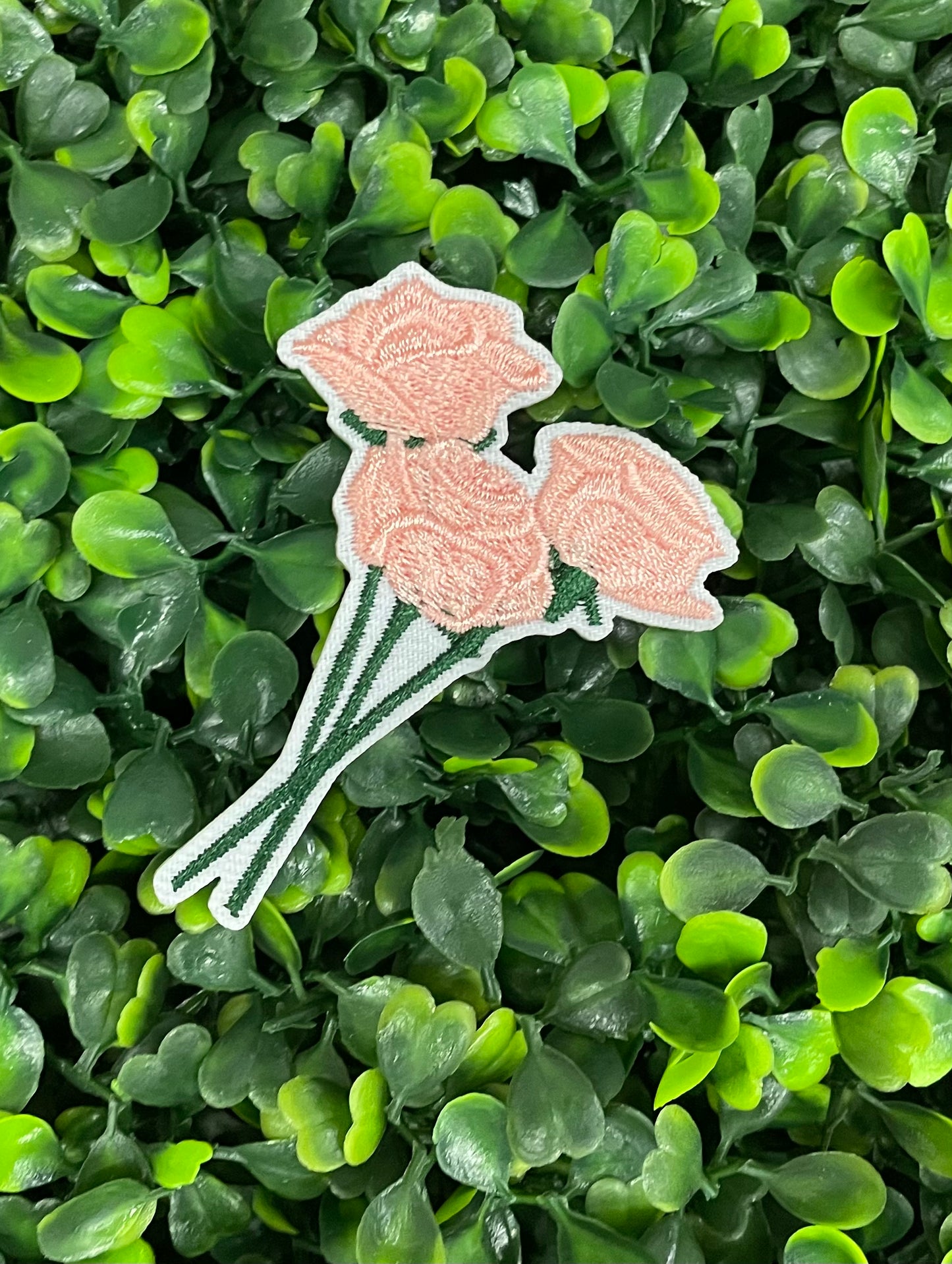 A hand holding an iron-on patch with embroidered pink roses attached to a green stem, set against a background of greenery.