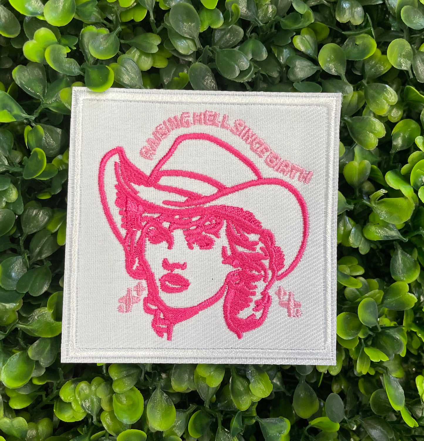 Embroidered patch with pink design and text "raising hell since birth" on a white background, placed on a green leafy backdrop