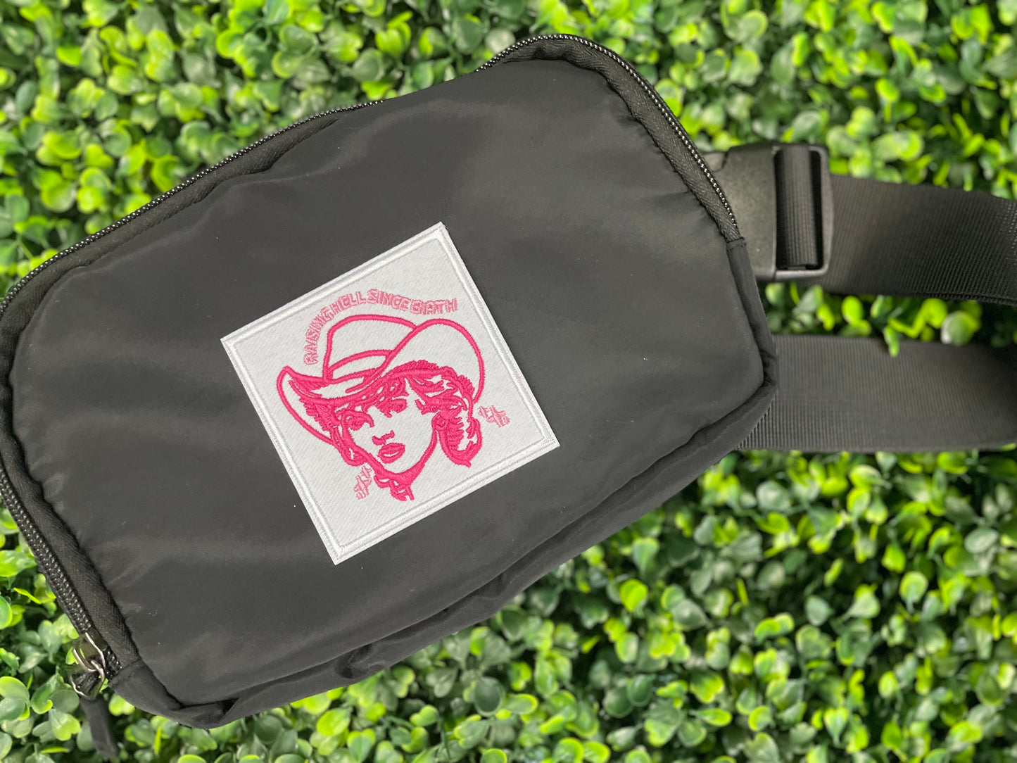 Embroidered patch with pink design and text "raising hell since birth" on a white background, placed on a black fanny pack