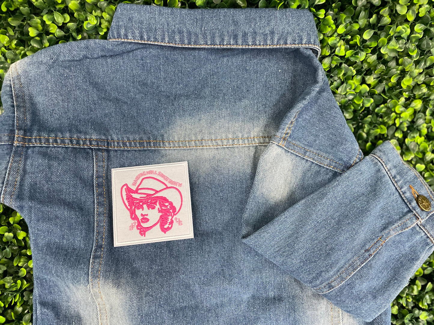 Embroidered patch with pink design and text "raising hell since birth" on a white background, placed on a children's denim jean jacket