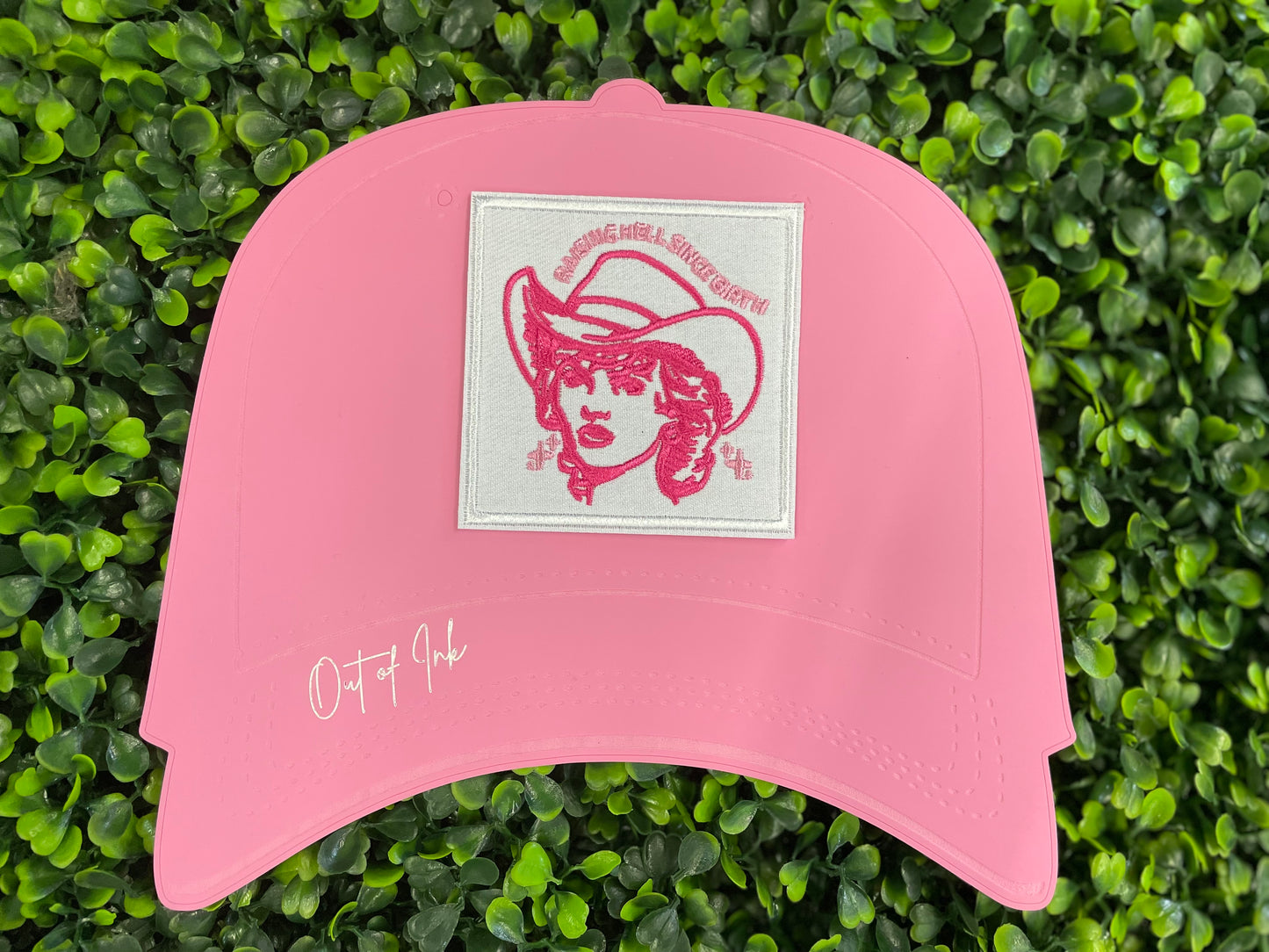 Embroidered patch with pink design and text "raising hell since birth" on a white background, placed on a pink hat graphic