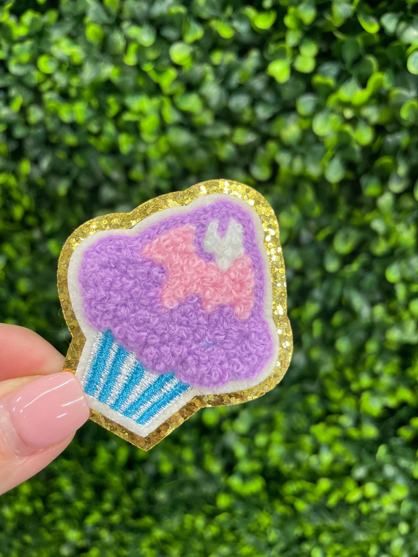 Decorative patch of a cupcake with pink and purple frosting, held against a green leafy background