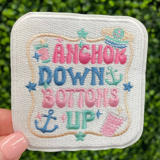 Embroidered patch with 'Anchors Down, Buttons Up' text held against a green leafy background