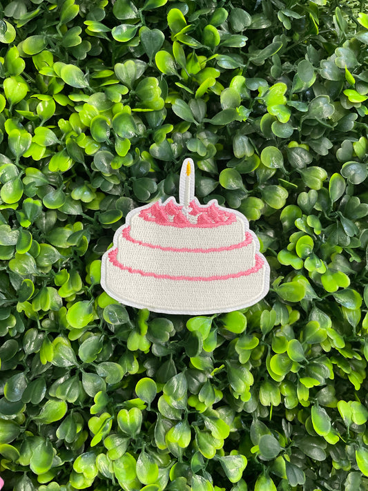 A small pink and white birthday cake embroidered patch placed on top of a green leafy background