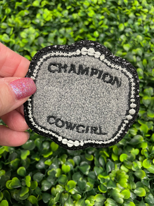 A close-up of an embroidered patch with the text 'Champion Cowgirl' held against a green leafy background.