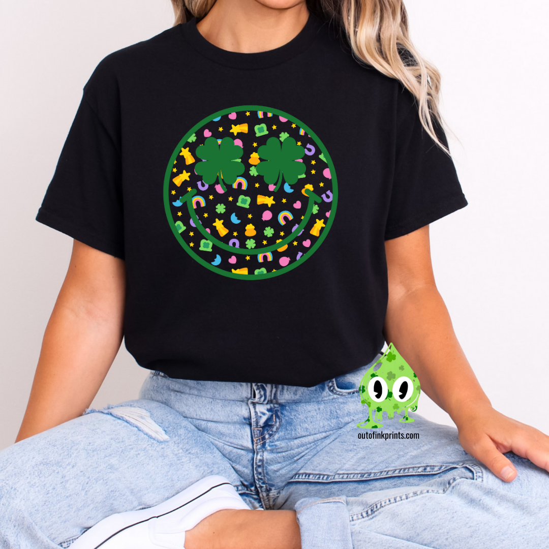 Front view of a black t-shirt with a smiley face design featuring clovers, celebrating St. Patrick's Day.