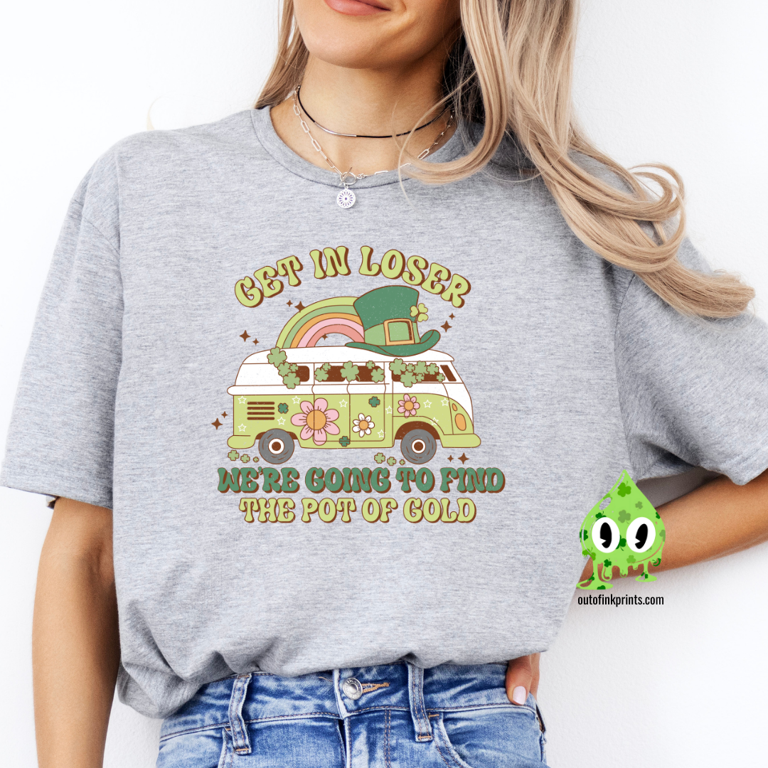 A woman wearing a gray t-shirt with the text 'GET IN LOSER ST. PATRICK'S' and a graphic of a pot of gold and a leprechaun.