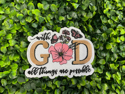 Embroidered patch butterflies and flowers and text stating "with God all things are possible", held against a green leafy background