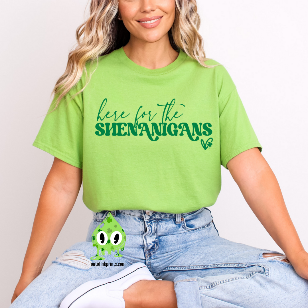 A person wearing a green t-shirt with the phrase 'here for the SHENANIGANS' printed in white, sitting on a bench.