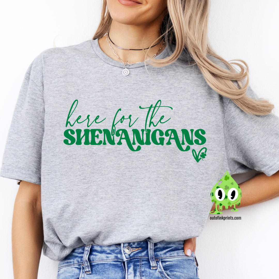 A person wearing a gray t-shirt with the phrase 'here for the SHENANIGANS' printed in white, sitting on a bench.
