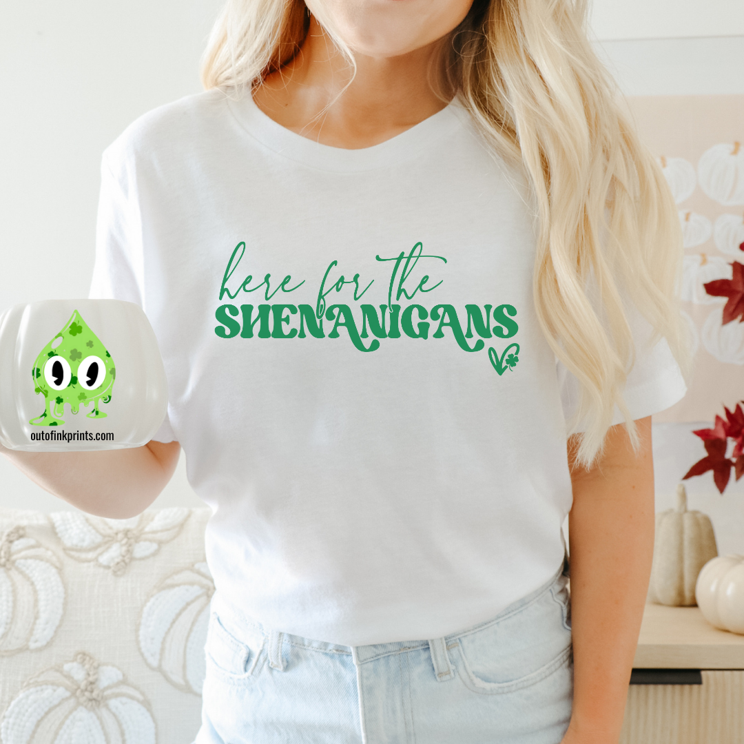 A person wearing a white t-shirt with the phrase 'here for the SHENANIGANS' printed in white, sitting on a bench.