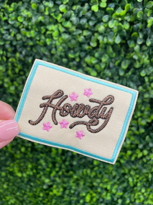 Embroidered patch with brown "Howdy" text on a tan background with a light blue border, placed against a green and leafy background