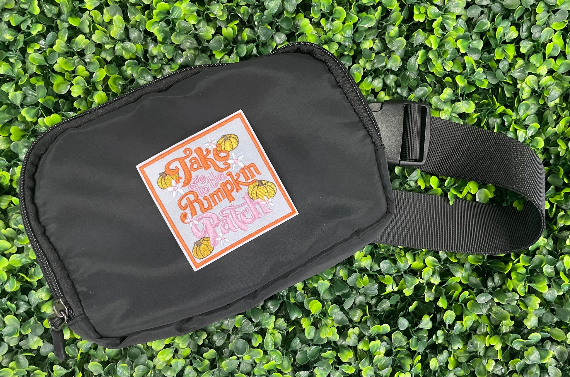 Embroidered patch featuring orange and pink text "Take me to the pumpkin patch" with pumpkin and flower designs around it, placed on a black fanny pack