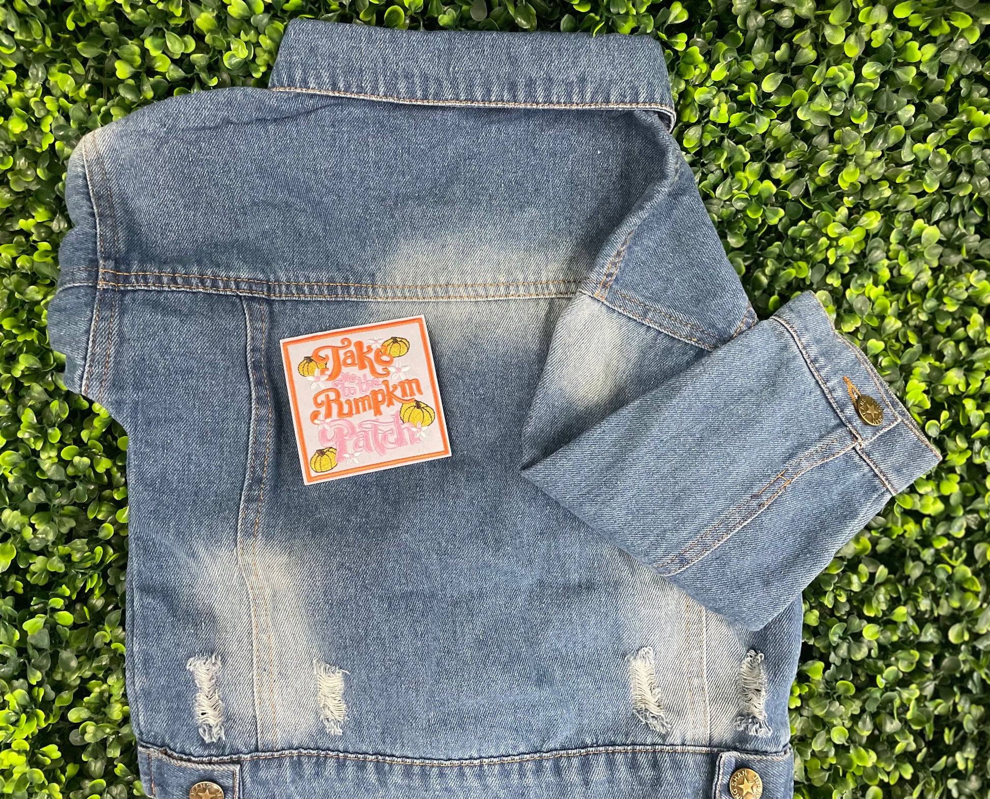 Embroidered patch featuring orange and pink text "Take me to the pumpkin patch" with pumpkin and flower designs around it, placed on a children's denim jean jacket