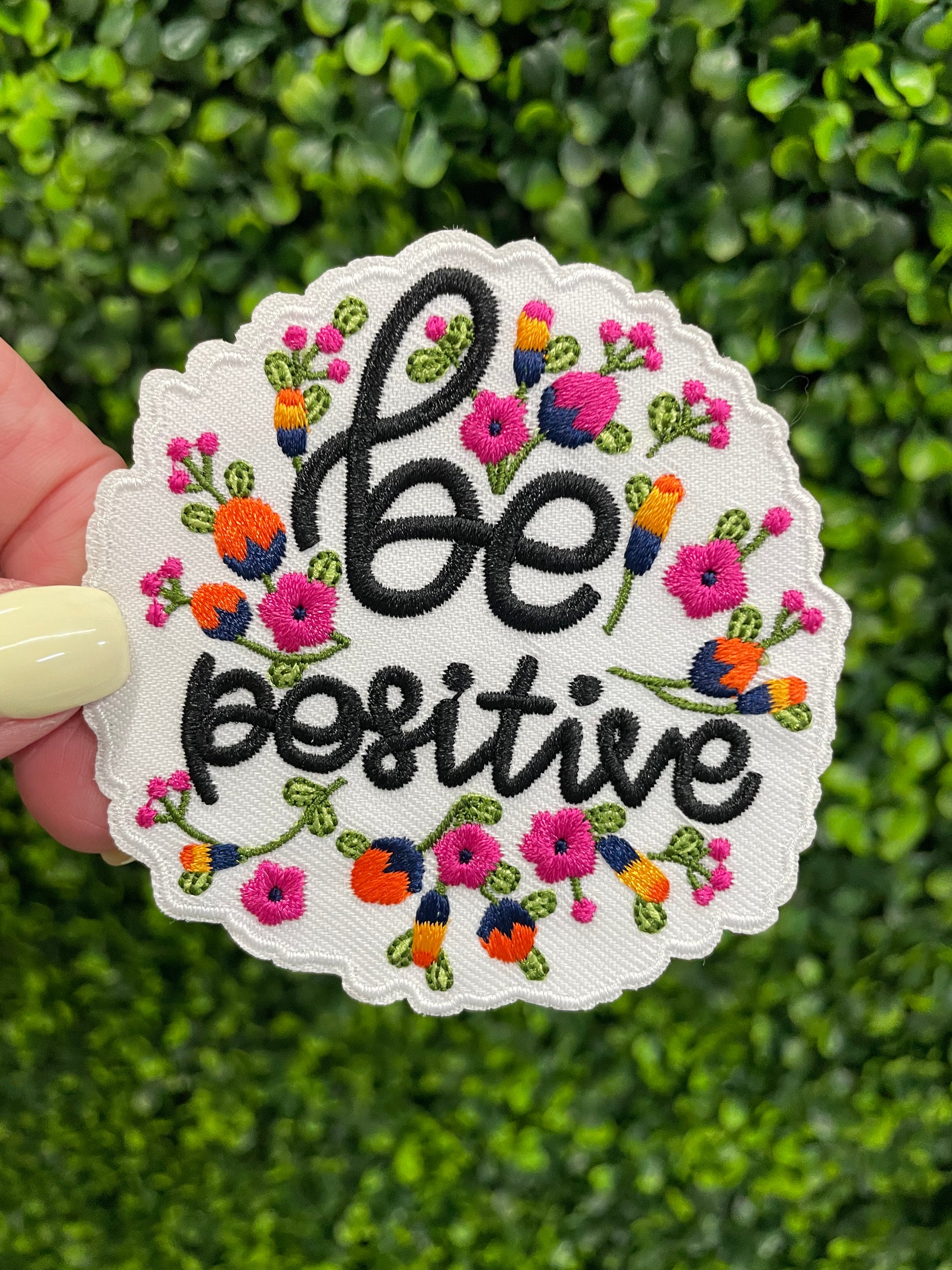 Embroidered patch with 'be positive' text and floral designs held against a green leafy background.