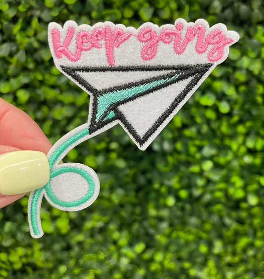 Embroidered patch with 'Keep going' text and paper airplane design held against a green leafy background